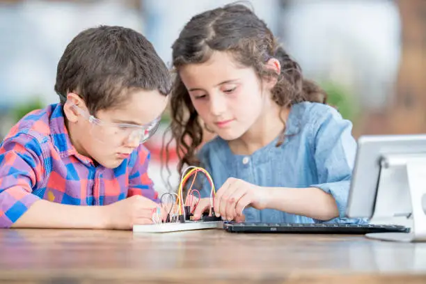 A young brother and sister are at school, working on a science project. They working with circuits and wires. They also have a tablet computer on a stand.