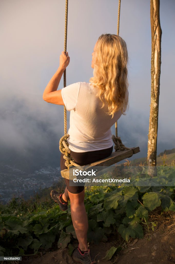 Woman steps off edge to swing into clouds She looks off to distant scene and volcano Banos Stock Photo
