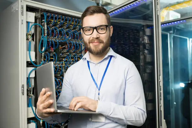 Photo of Portrait of content confident bearded highly professional IT engineer with badge on neck using laptop in network server room