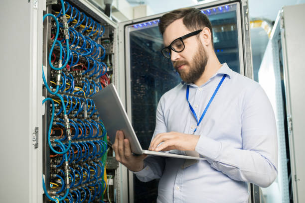 istock Concentrated handsome technical specialist in glasses managing supercomputer via laptop and standing at server rack cabinet in modern storage room 964442174