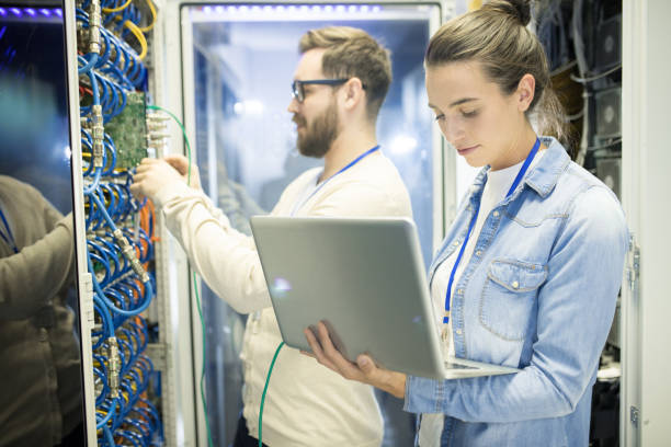 istock Serious professional female technician holding research on laptop while thinking about efficiency of supercomputer inside of server cabinet 964442416