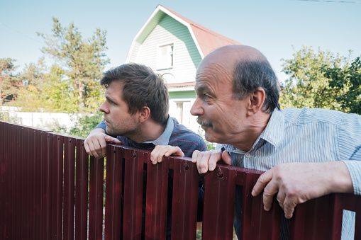 two caucasian men carefully watching over the fence. Concept of curious neighbors and private life