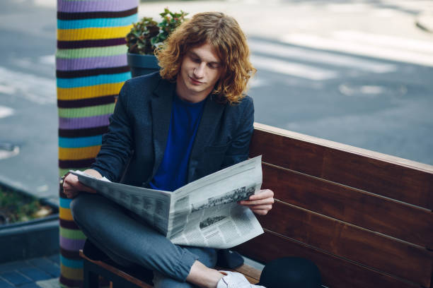 istock Red haired hipster man sitting on bench reading newspaper 964486974