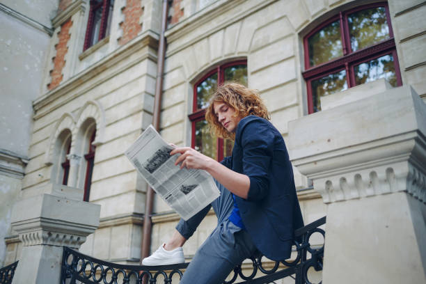 istock Young reddish man reading newspaper near old style building 964487104