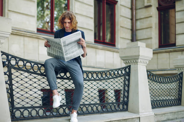 istock Young reddish man reading newspaper near old style building 964487262