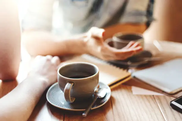 Photo of Friends talking at cafe table during coffee break. Unrecognizable male and female colleagues discussing business issues, focus on coffee cup with saucer and teaspoon