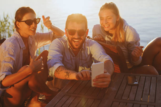 Two young woman and man using smartphone by the river and smiling