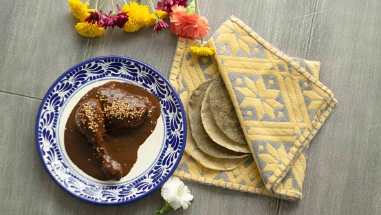 A Mexican traditional plate with two pieces of chicken covered in the Mexican sauce mole. The table is wooden, there are four corn tortillas in a yellow table cloth to keep them warm. There are also some flowers on top of the table.