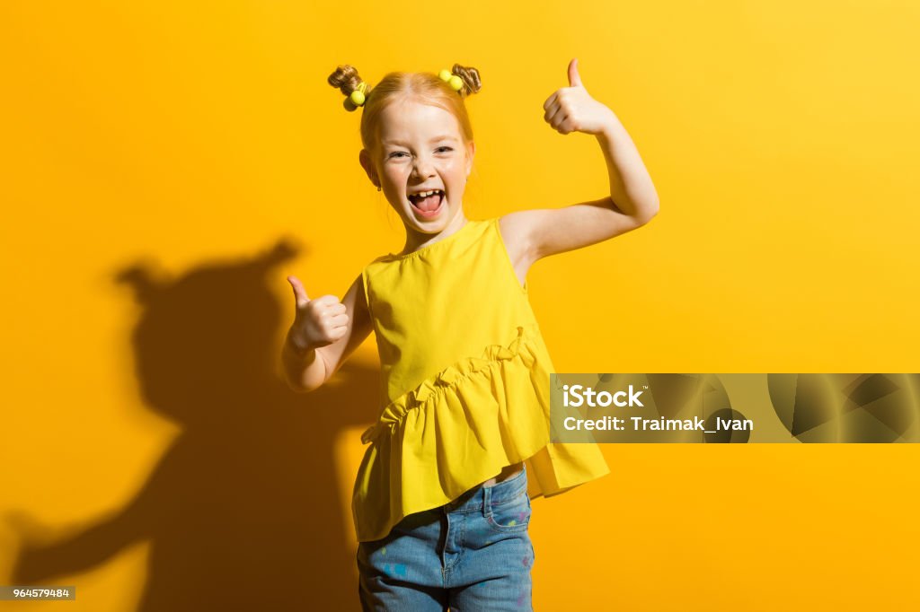 Girl with red hair on a yellow background. The girl laughs and shows the class sign. Portrait of a beautiful girl in a yellow blouse and blue jeans. Child Stock Photo