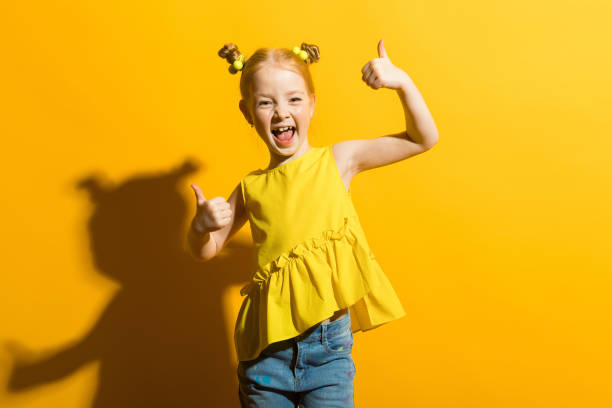 Girl with red hair on a yellow background. The girl laughs and shows the class sign. Portrait of a beautiful girl in a yellow blouse and blue jeans. cute girl stock pictures, royalty-free photos & images