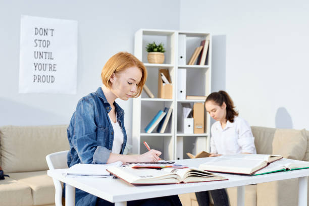 Serious concentrated attractive redhead student girl making notes in diary while planning work, her busy female friend sitting in armchair and reading book in background Students focusing on new information exam routine preparation education stock pictures, royalty-free photos & images