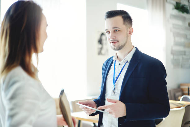 istock Confident young businessman with tablet computer in his hands and accreditation badge around his neck talking with unrecognizable female colleague at business event 964701016