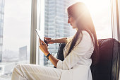 Successful woman wearing suit sitting on armchair using tablet computer at her loft apartment in city center