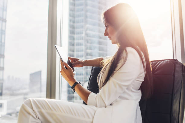 Successful woman wearing suit sitting on armchair using tablet computer at her loft apartment in city center Successful woman wearing suit sitting on armchair using tablet computer at her loft apartment in city center. well dressed stock pictures, royalty-free photos & images