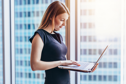 Portrait of young woman working holding laptop standing against panoramic window with city view