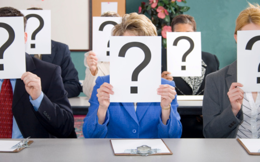 Group of business woman and men sitting at table with question marks held in front of face.