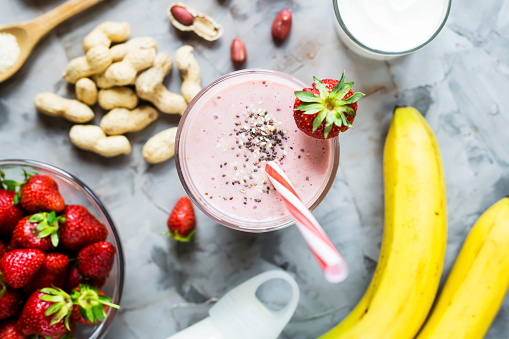 A glass of strawberry banana smoothie among the ingredients for its cooking - strawberries, bananas, milk, yogurt, chia seeds, sesame and peanuts. Healthy eating breakfast. Top view, flat lay