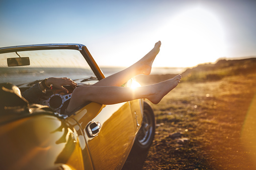 Woman on road trip relaxing at beach lying down in retro convertible car at sunset