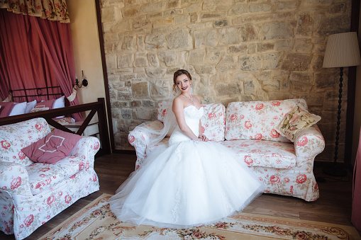 Una chica en un vestido blanco ajustado se sienta en el borde del sofá en su cuarto. Una casa de madera y ambiente acogedor hacen la novia sonriendo photo