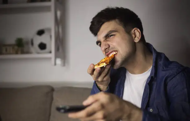 Photo of Man eating pizza and holding remote control