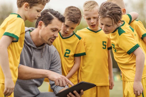 una squadra di calcio per ragazzi che guarda le tattiche su un tablet con il loro allenatore maschio durante una partita - strategy coach soccer digital tablet foto e immagini stock