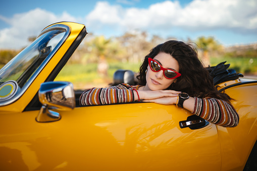 Young fashionable hipster woman with retro sunglasses relaxing in vintage cabriolet car on summer vacations