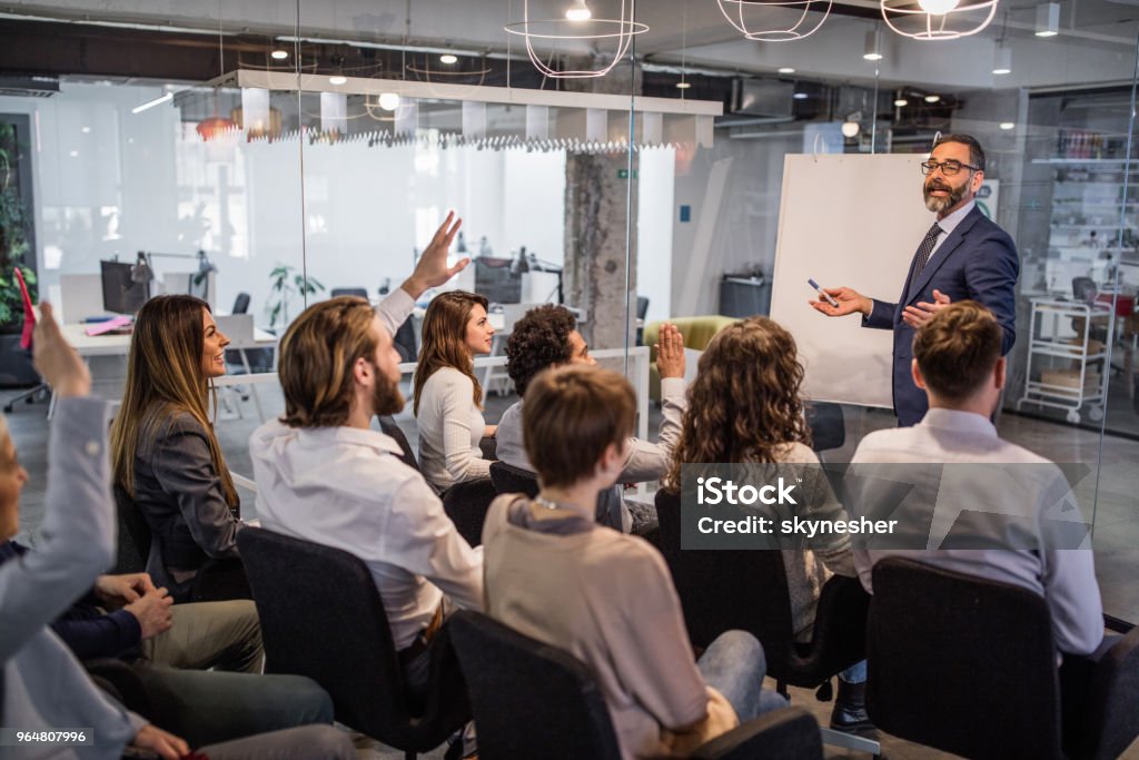 Happy CEO giving his team a business presentation in a board room. Mature businessman talking to large group of people on a business presentation in a board room. Some of them are raising their hands. Education Training Class Stock Photo