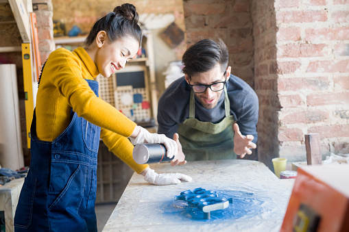 Portrait of two modern artists looking excited while spray painting wood while working in carpenters workshop, focus on smiling woman in foreground