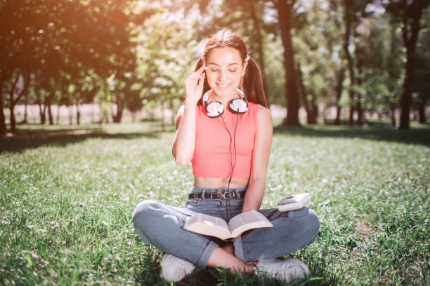 istock Smart student is sitting on grass in park and reading a book. She is sitting with her legs crossed. Girl is holding one hand close to head and smiling 964842814