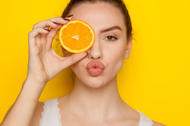 Young woman posing with slice of orange on her face on yellow background Young woman posing with slice of orange on her face on yellow background puckering stock pictures, royalty-free photos & images