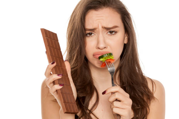 istock a young beautiful woman is tempted by chocolate and vegetable on white background 964883528