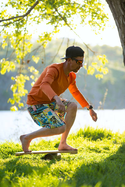 Bearded man is training on the balance board and having fun on a green meadow next to river on sunny evening. Back light.