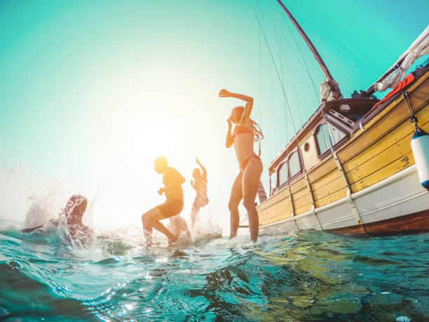 szczęśliwi przyjaciele nurkujący z żaglówki do morza - młodzi ludzie skaczą do oceanu w letniej wycieczce - wakacje, młodzież i zabawa - główny nacisk na sylwetkę facetów - zniekształcenie obiektywu rybie oko - sunset yacht luxury sailboat zdjęcia i obrazy z banku zdjęć