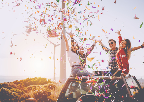 Portrait group of cool teen hipster friends throwing colorful confetti during partying and enjoy outdoors. They are smiling and looking at camera posing on car at natural wind farm area on sunny summer day while carefree road trip