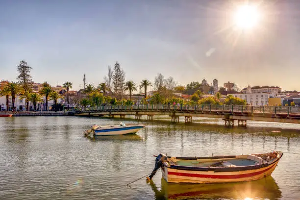 Beautiful post card, with boats on serene Gilao river, and the sun shining over picturesque Tavira, a popular tourist destination in southern Portugal.