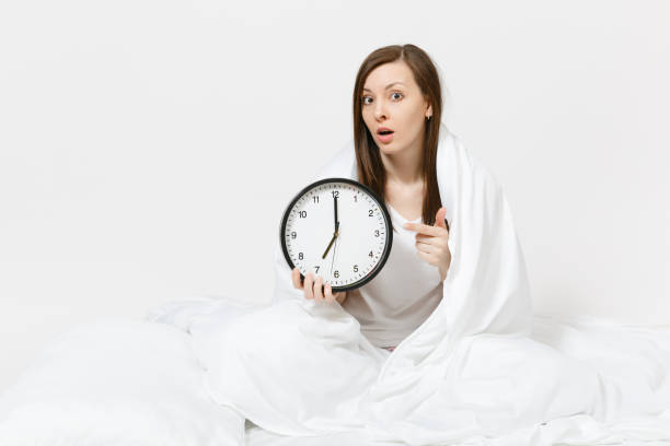 istock Young woman sitting in bed with round clock, white sheet, pillow, wrapping in blanket on white background. Beauty female wake up early in morning, spending time in room. Rest, relax, good mood concept 964976594