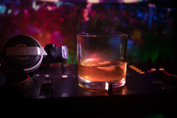 istock Glass with whisky with ice cube inside on dj controller at nightclub. Dj Console with club drink at music party in nightclub with disco lights. 964985422