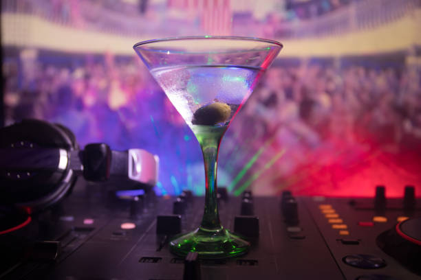 istock Glass with martini with olive inside on dj controller in night club. Dj Console with club drink at music party in nightclub with disco lights. Close up view 964985456