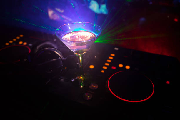 istock Glass with martini with olive inside on dj controller in night club. Dj Console with club drink at music party in nightclub with disco lights. Close up view 964985472