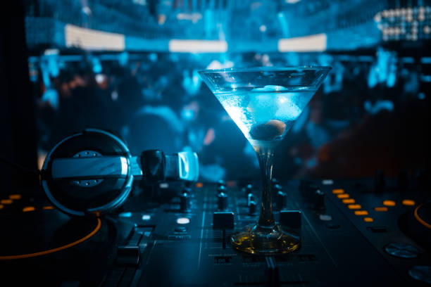 istock Glass with martini with olive inside on dj controller in night club. Dj Console with club drink at music party in nightclub with disco lights. Close up view 964985500