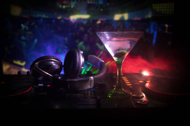 istock Glass with martini with olive inside on dj controller in night club. Dj Console with club drink at music party in nightclub with disco lights. Close up view 964985550