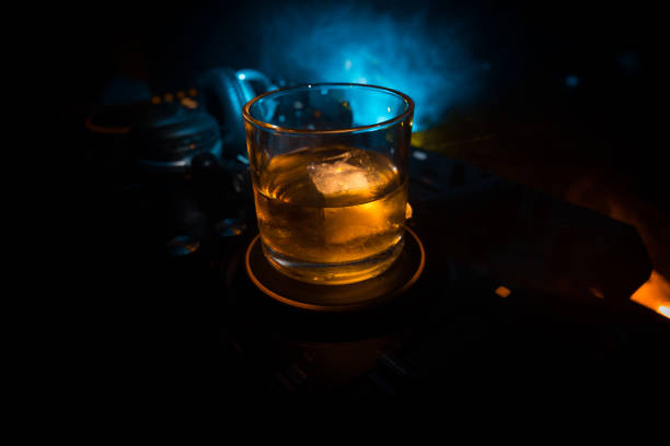 Glass with whisky with ice cube inside on dj controller at nightclub. Dj Console with club drink at music party in nightclub with disco lights. Selective focus