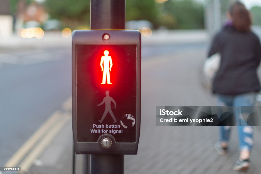 red stop symbol or don't cross sign push button wait for signal traffic light control with bright stop man illuminated at pedestrian crossing and woman walking past Amber Stock Photo
