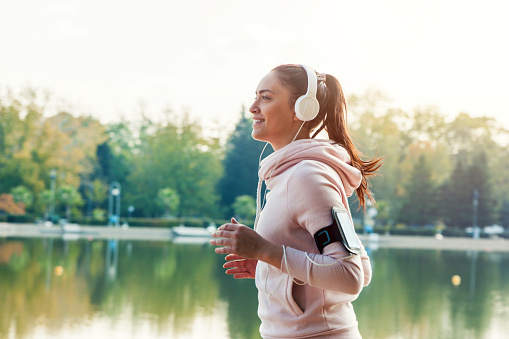 A young woman with a ponytail is wearing sports clothing and running along a body of water in a park. She is listening to music with headphones and her smartphone is in an armband. With copy space.
