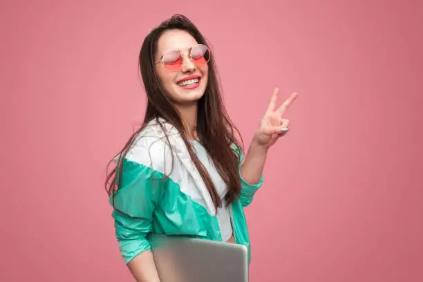 Trendy millennial model in sunglasses holding modern laptop and showing two fingers with smile on pink.