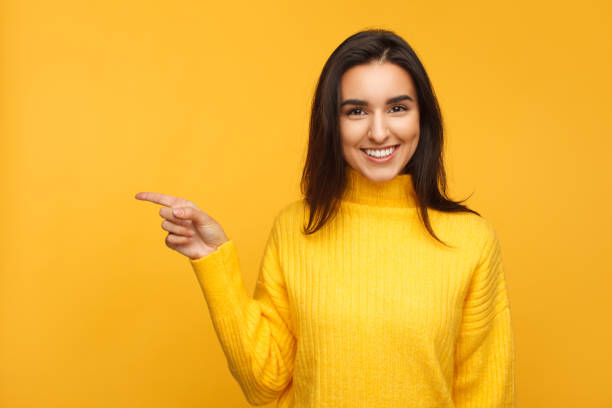 Pretty brunette directing to left Stylish smiling girl in yellow sweater pointing away standing on yellow backdrop. left handed stock pictures, royalty-free photos & images