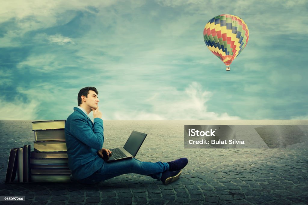 educational process Thinking student sitting on a pavement floor lean on a pile of books, using laptop and a hot air balloon flying. Modern education concept. New technology replace old methods in educational process. Book Stock Photo