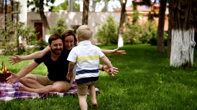 Family idyll. Happy child running to his parents in a blurred perspective. Close up of young couple hugging his little blonde son while sitting on the grass, plaid. Picnic in the park. Slow motion