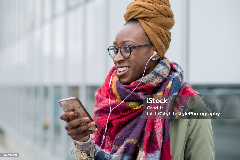 Standing Downtown A beautiful woman of African descent is standing next to a building downtown. She is dressed fashionably in trendy clothing. She smiles and uses her smartphone and earbuds to listen to music. 20-29 Years Stock Photo