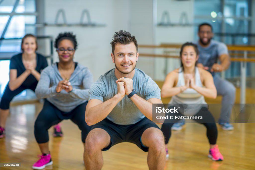 Doing Squats A group of adults are taking a fitness class together. They are arranged in rows and are smiling at the camera while doing squats. Exercise Class Stock Photo